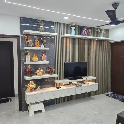 TV wall in beige fluted slats with a black marble-look slab bay carrying four staggered white display shelves of brass deity idols and red rose arrangements