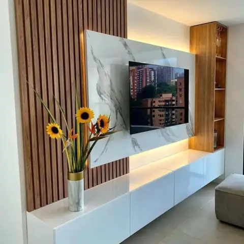 TV wall where a white marble-veined panel is set proud of vertical teak fluting and backlit on all four sides, above a glossy white floating console