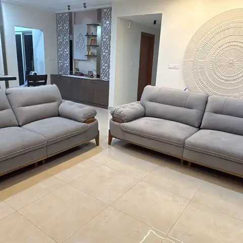 Two grey fabric sofas on teak legs face each other across glossy beige tile, with a large carved radial sunburst medallion on the wall behind and a