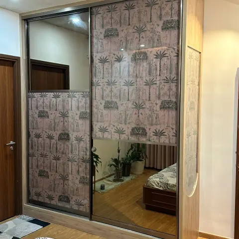 Two-shutter sliding wardrobe faced in blush pichhwai-print glass and mirror, oak-laminate framing, standing beside the bed in a fan-lit bedroom.
