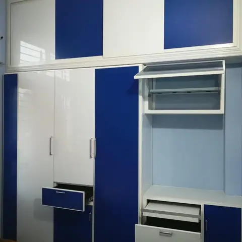 Two-tone blue and white glossy wardrobe wall with open shelving and pull-out drawers in a child's bedroom, wooden bed frame visible in the corner.