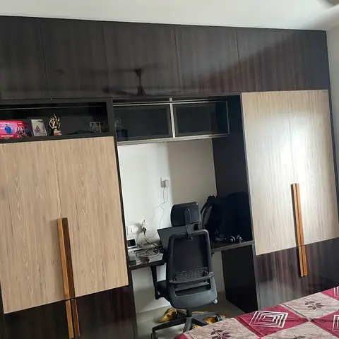 Two-tone brown and light-oak laminate wardrobe wall with a built-in study desk and office chair, personal trophies displayed on the open shelf above.