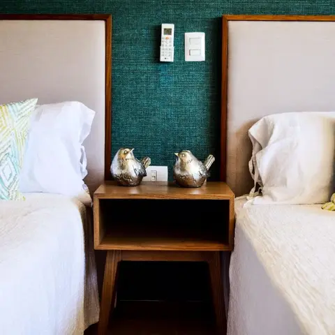 Two twin beds with beige upholstered headboards flanking a small wood nightstand against a teal woven-textured wallpaper wall