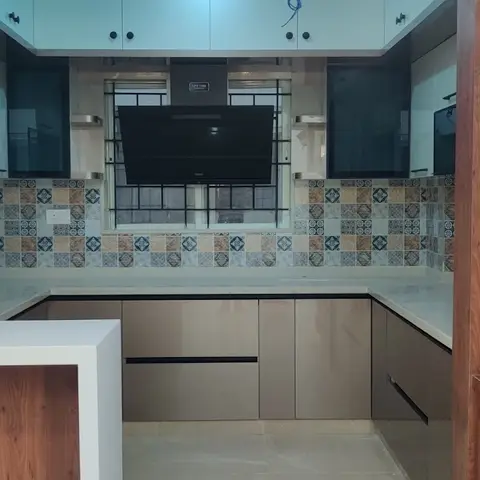 U-shaped kitchen with a patchwork mosaic-tile backsplash, a wall-mounted TV above the hob, and a carved wood pooja cabinet with brass bells visible alongside.