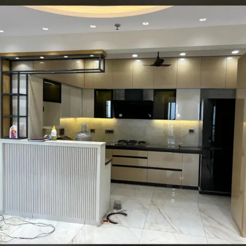 U-shaped modular kitchen mid-handover with high-gloss beige shutters, a fluted-panel breakfast counter, marble-look backsplash and a round cove-lit ceiling