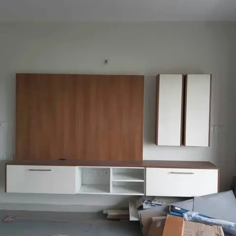 Under-construction living room wall with a walnut-panelled TV backdrop and matching white overhead storage cabinets, packaging debris still on the floor.