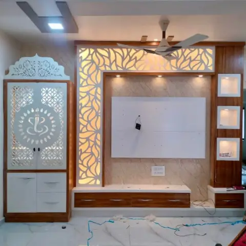 Under-construction TV feature wall with a backlit floral CNC-jaali panel and marble-look dado, alongside a white pooja cabinet with a Ganesh-motif jaali door.
