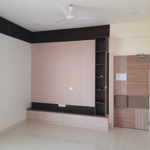 Unfurnished living room with a dusty-pink panelled TV wall unit featuring open shelving and a dark ridged trim band, opposite a patterned laminate door.