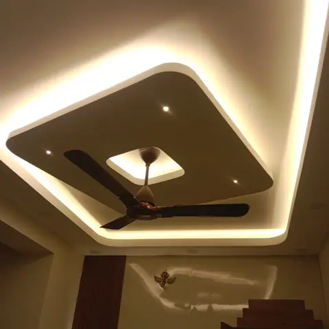 Upward view of a diamond-shaped cove-lit false ceiling with a ceiling fan, above a wooden pooja mandir with beaded toran hangings visible below.