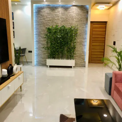 View across a glossy tiled living room to a stone-strip clad feature wall lit blue-white from behind a low white planter of tall bamboo