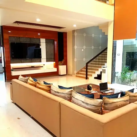 Villa living room seen over a big beige L-sofa toward a TV wall of glossy red-brown veneer framing dark stone panels above a lit floating console
