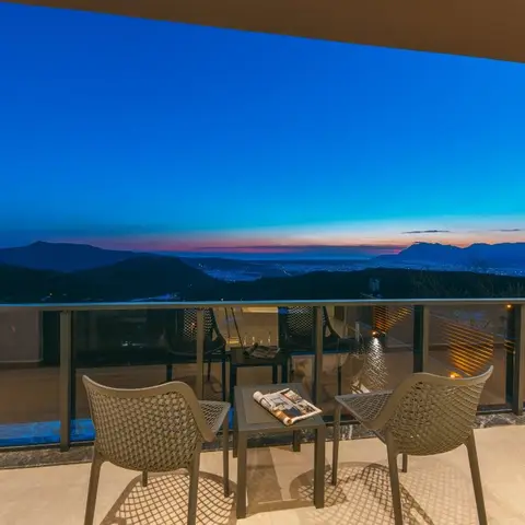 Villa terrace at dusk with mesh-weave outdoor chairs and a small side table, overlooking a mountain and city skyline through a glass balustrade.