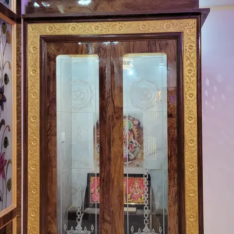 Walk-in pooja room shut with twin etched-glass doors carrying Om, diya and swastik motifs, framed in gold floral carved beading over burl-veneer