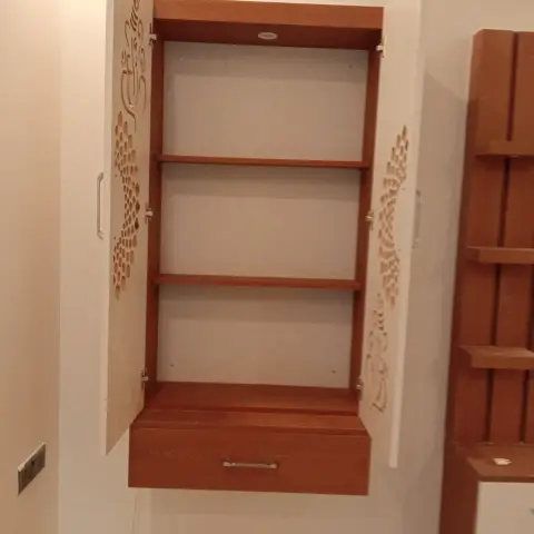 Wall-hung pooja cabinet with both white CNC-cut shutters swung open - a Ganesha figure cut into one leaf - showing three teak-laminate shelves