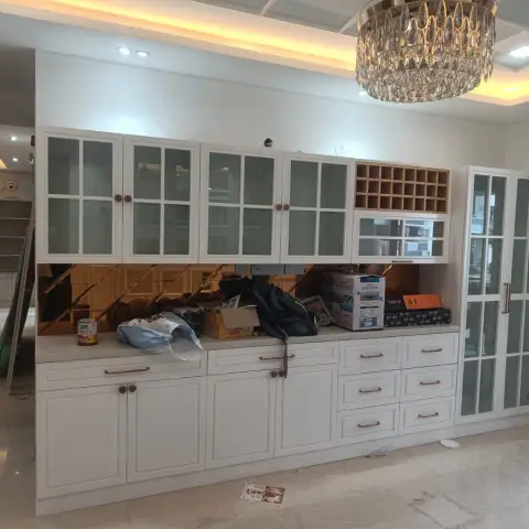 Wall-length white shaker crockery and bar run with a rose-gold mirror backsplash and honeycomb wine rack, under a coffered cove ceiling and crystal chandelier