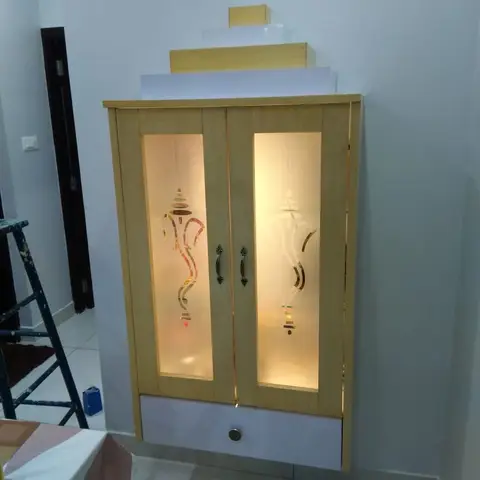 Wall-mounted pooja cabinet with backlit frosted glass doors etched with a Ganesha motif, a tiered wood pelmet top, and a lower drawer with a brass knob.
