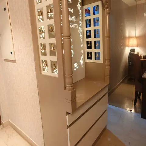 Wall-mounted pooja unit in taupe lacquer with a carved jaali pediment, backlit bell niches on either side, and drawer storage below.