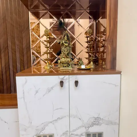 Wall-mounted pooja unit with a diamond-pattern mirror backsplash, marble-effect laminate cabinet doors, and brass idols and diyas on the wood-laminate shelf.