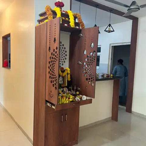Wall-mounted pooja unit with jaali-cut doors open to reveal deities and marigold garlands, kitchen counter visible through the archway beyond
