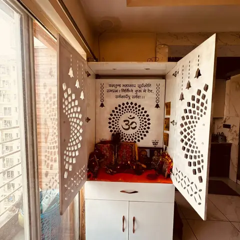 Wall-mounted pooja unit with perforated Om-motif jaali doors, a white laminate cabinet base and deities on a red cloth