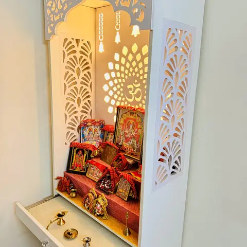 Wall-mounted white pooja unit with a CNC-cut jaali screen backlit in warm LED, a tiered shelf holding framed deities, and a pull-out drawer for diyas.