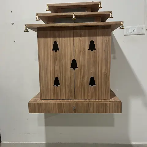 Wall-mounted wooden pooja unit with a tiered canopy roof, bell-motif cutout doors, and hanging brass bells.