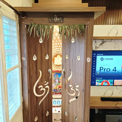 Wall-mounted wooden pooja unit with a stepped temple-style pediment, gold Om-carved bifold doors hung with bells and a fresh mango-leaf toran