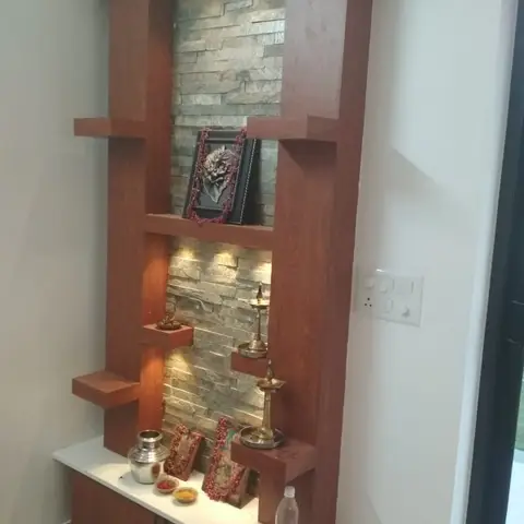 Wall-mounted wooden pooja unit with staggered display shelves, a stacked stone-cladding backdrop, and brass lamps and kalash set out for worship.