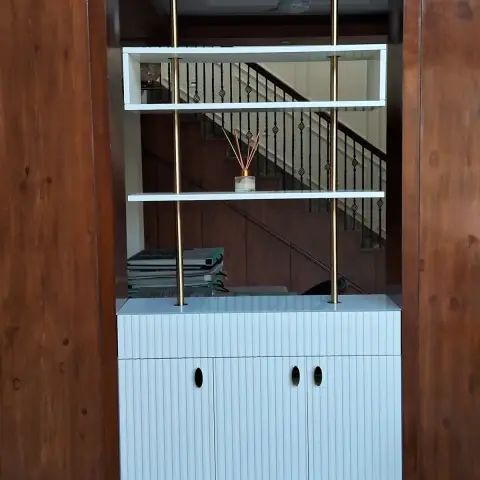 Wall-panelled display cabinet with fluted white cabinetry, brass support rods and glass shelving set into a dark wood-veneer wall