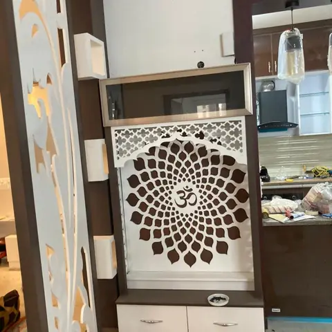 Wall-recessed pooja unit with a white CNC-cut jaali shutter forming a lotus and Om motif over a mirrored glass cabinet, beside a matching wall jaali partition.