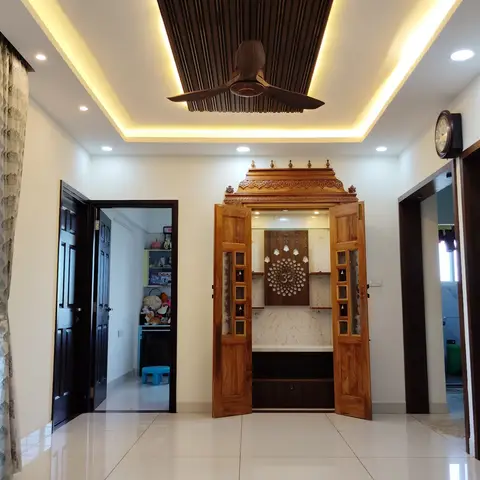 Wall-recessed pooja unit with an intricately carved teak door frame, a brass Om motif jaali panel, and a marble backdrop under a cove-lit ceiling.