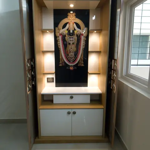 Wardrobe-style pooja unit with an internal backlit deity photo, brass door bells on both shutters, and a white two-door cabinet base.