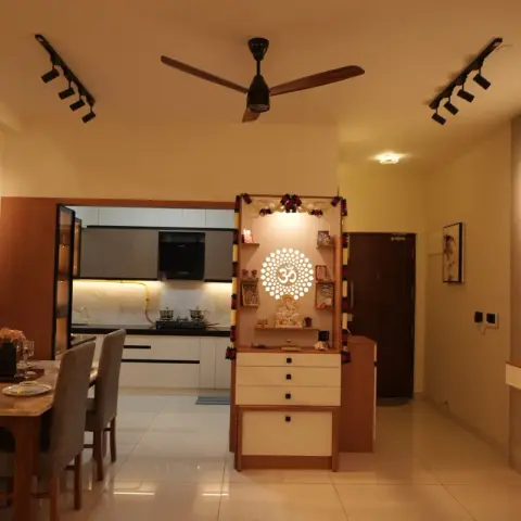 Warm evening view across an open plan where a freestanding pooja unit with a backlit Om cut-out and marigold torans splits the dining table from the kitchen