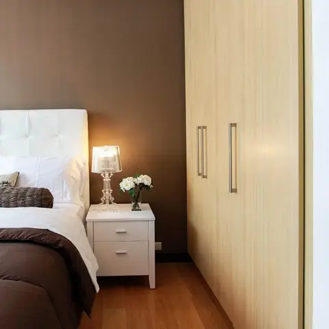 White button-tufted leather headboard against a taupe accent wall, with a pale maple-veneer wardrobe run alongside and a ghost-style acrylic table lamp on a