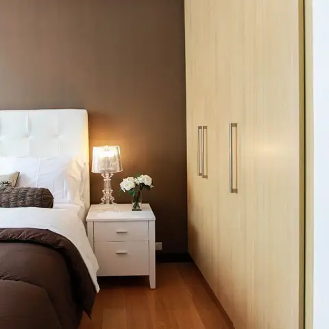 White button-tufted leather headboard against a chocolate-brown accent wall, with a clear ribbed table lamp and white roses on a two-drawer white nightstand