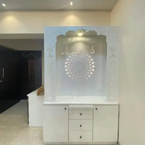 White CNC-carved pooja unit with a backlit Om lotus jaali panel and storage drawers below, set beneath a cove-lit ceiling.