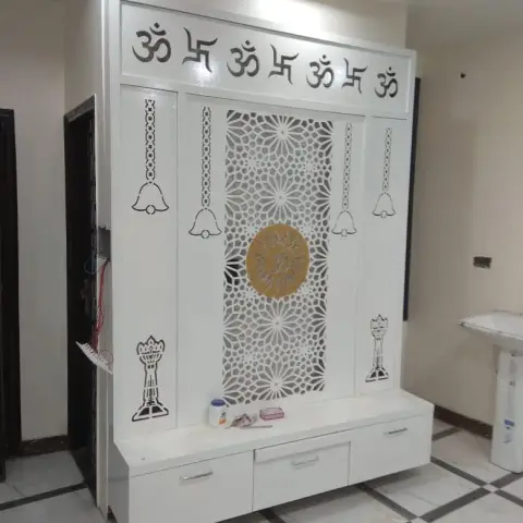 White CNC-jaali pooja unit with Om and swastik motifs and hanging bell cutouts, over drawer storage, set against a black-bordered marble floor