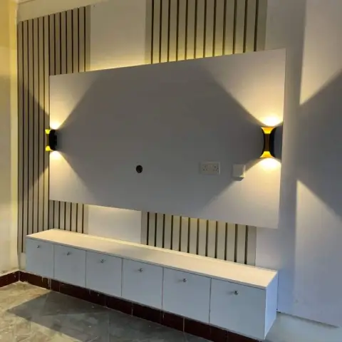 White fluted-panel TV feature wall with a backlit rectangular frame and twin black-and-gold up-down wall sconces above a floating white TV console.