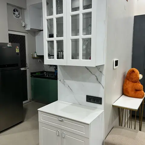 White glass-front crockery unit with a marble backsplash and quartz-top base cabinet, positioned at the entrance to an adjoining green modular kitchen.