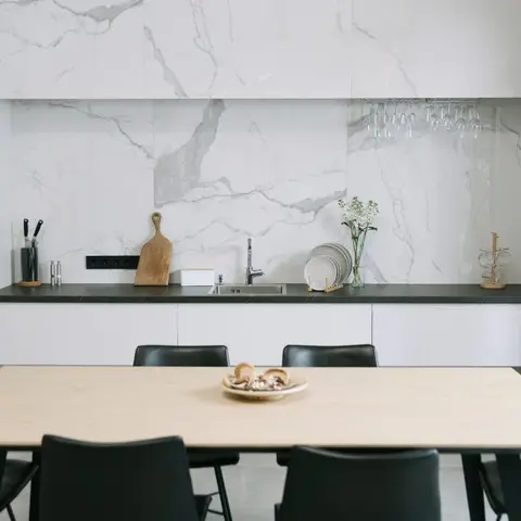 White handleless galley kitchen with a marble-look slab backsplash and black countertop, seen behind a light wood dining table with black leather chairs