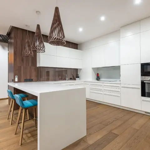 White handleless kitchen with a walnut wood-clad hood wall, three geometric woven-wood pendant lights over a waterfall-edge island and teal bar stools