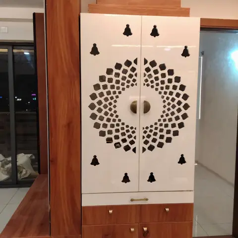 White high-gloss laminate pooja unit with laser-cut bell-motif jaali doors and wood-grain laminate drawer base, set against a foyer wall at night.