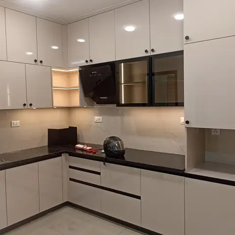 White high-gloss modular kitchen with a black stone countertop, a glass-front display cabinet, and a warm-lit open corner shelf