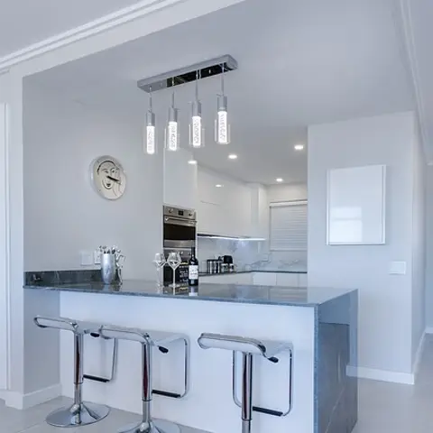 White kitchen breakfast counter with three chrome swivel bar stools under a row of cylindrical pendant lights