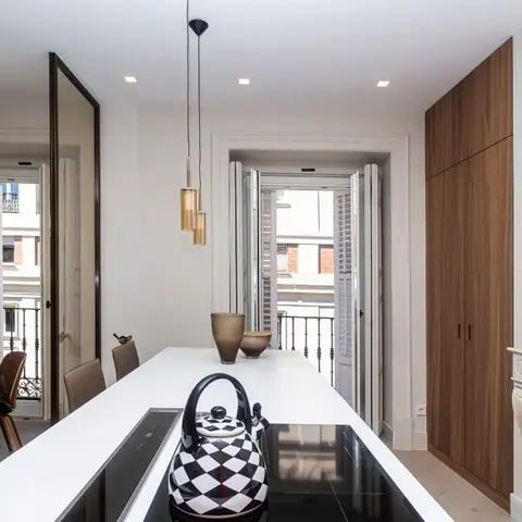 White kitchen island with induction hob facing an open balcony door, tall walnut pantry cabinets, and a marble fireplace mantel in the corner