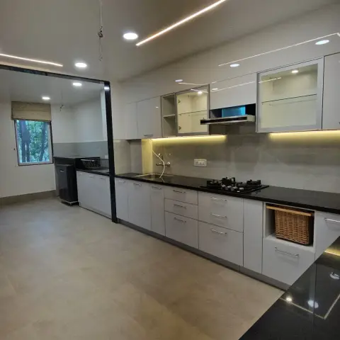 White L-shaped kitchen with a black granite counter and hob, glass-front upper cabinets, and an angular cove-lit false ceiling.