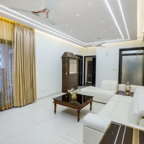 White leather L-sectional and a turned-leg wooden coffee table with a black glass top sit under a false ceiling cut by long angular linear-profile lights