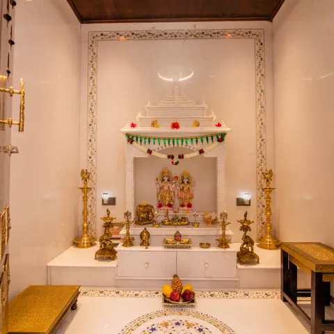 White marble pooja room with a carved temple-style mandir alcove housing deity idols, brass diyas and elephant statues either side