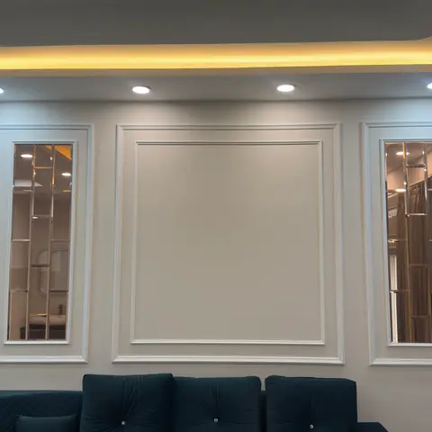 White panel-moulded feature wall with two mirrored inset panels flanking a plain panel, cove-lit ceiling above a teal tufted velvet sofa.