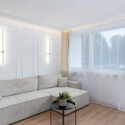 White panel-moulded living room corner with vertical LED wall lights flanking a beige corduroy sectional and sheer-curtained balcony door